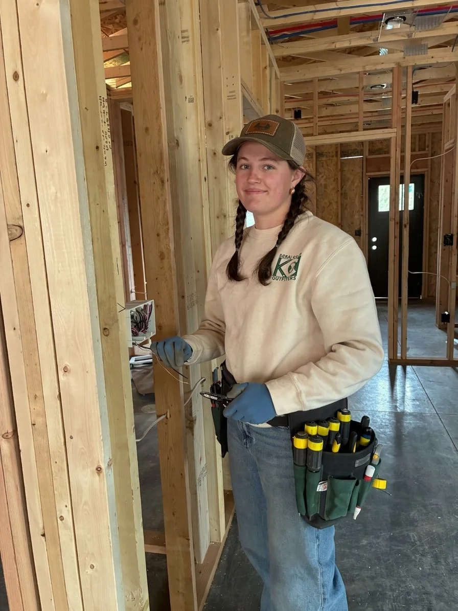 Electrician installing rough-in wiring and a main electrical panel inside the framed walls of a new residential home under construction in rural Custer County, Colorado. Open stud framing and mountain terrain are visible through an unfinished window opening.